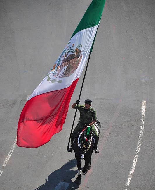 Мексиканский солдат верхом на коне скачет с флагом Мексики.