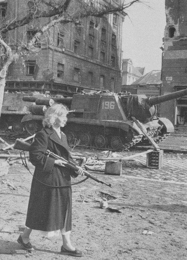 Девушка-повстанец с винтовкой идет мимо брошенной САУ ИСУ-152 во время Венгерского восстания, Будапешт, 1956 г.