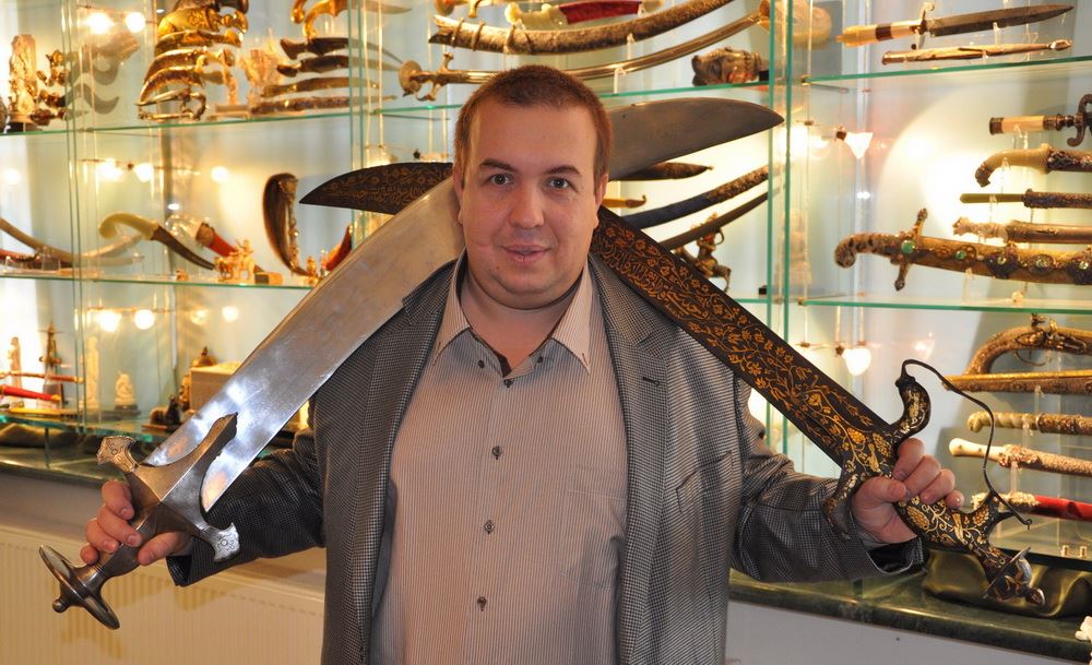 Коллекционер с двумя большими тальварами на фоне остальной коллекции восточного оружия, 2022 г.
